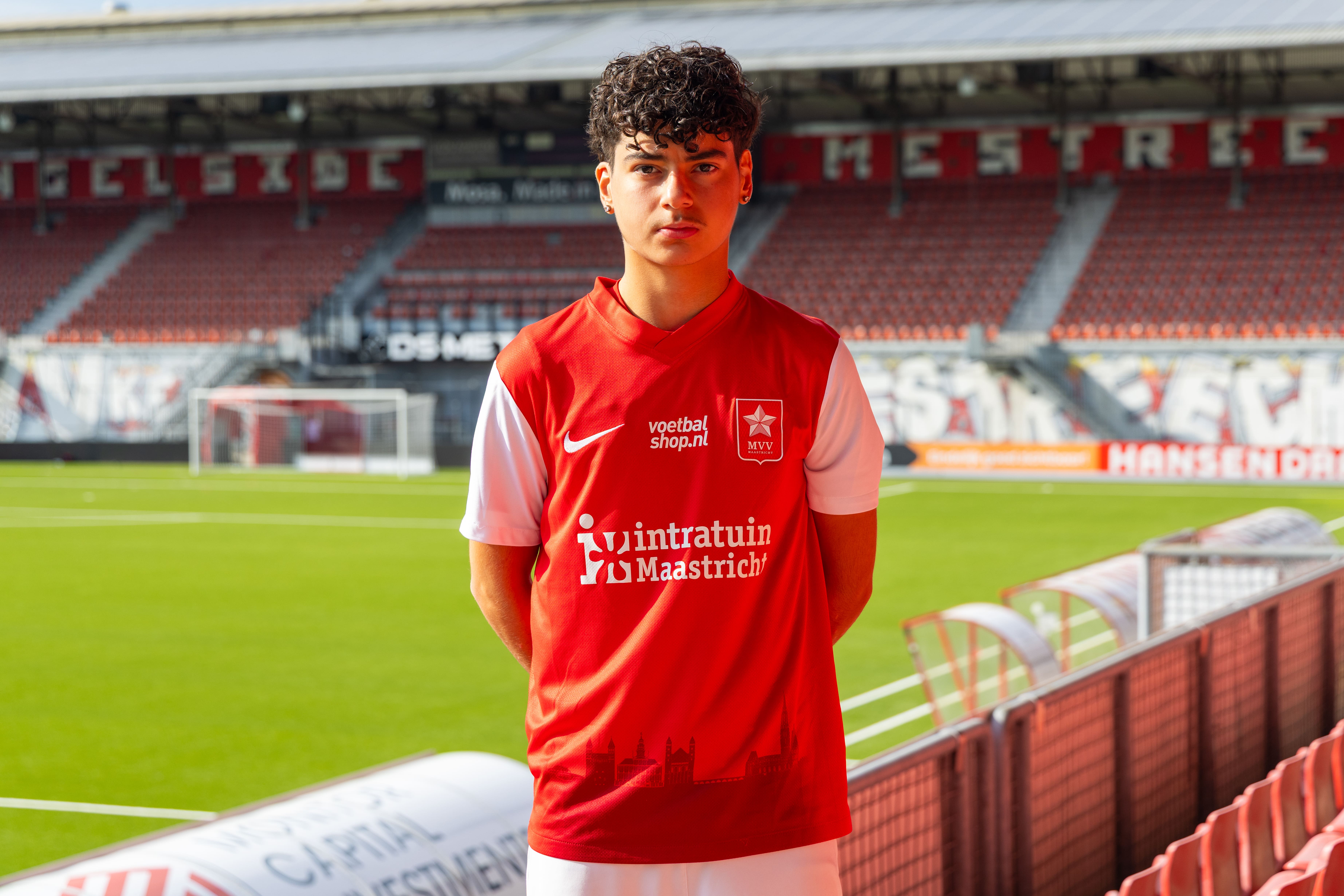 Young male soccer player in red and white MVV Maastricht jersey standing in an empty stadium.
