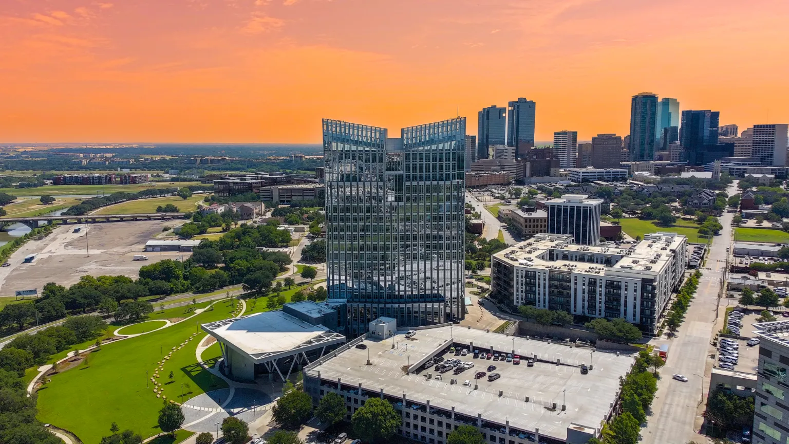 Aerial view of a cityscape at sunset, featuring a reflective glass skyscraper in the center. Surrounding buildings and green spaces create a modern urban feel.