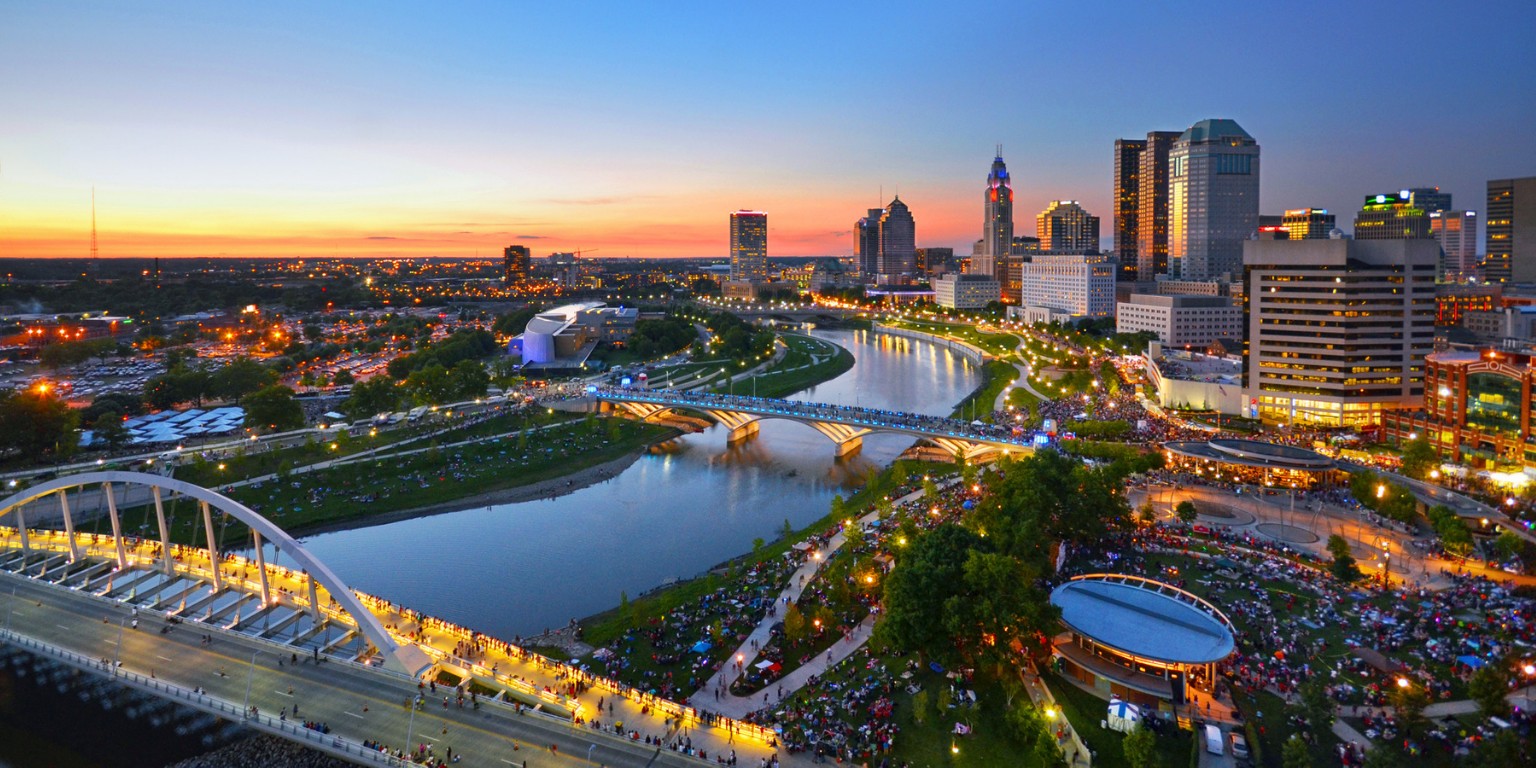 A vibrant cityscape of Columbus, Ohio, at dusk. Skyscrapers and buildings surround a winding river with illuminated bridges, set against a colorful sunset sky.