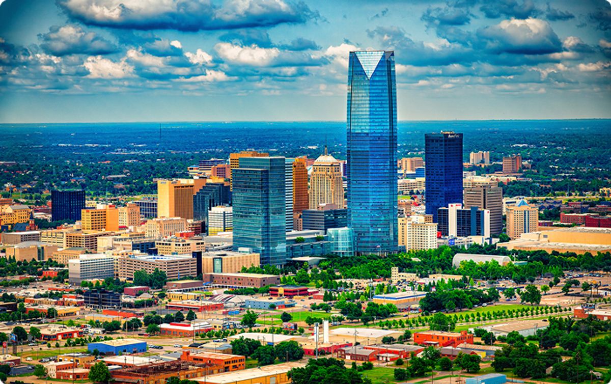 A vibrant cityscape featuring skyscrapers under a partly cloudy sky, with a prominent glass tower in the center, surrounded by lush greenery.