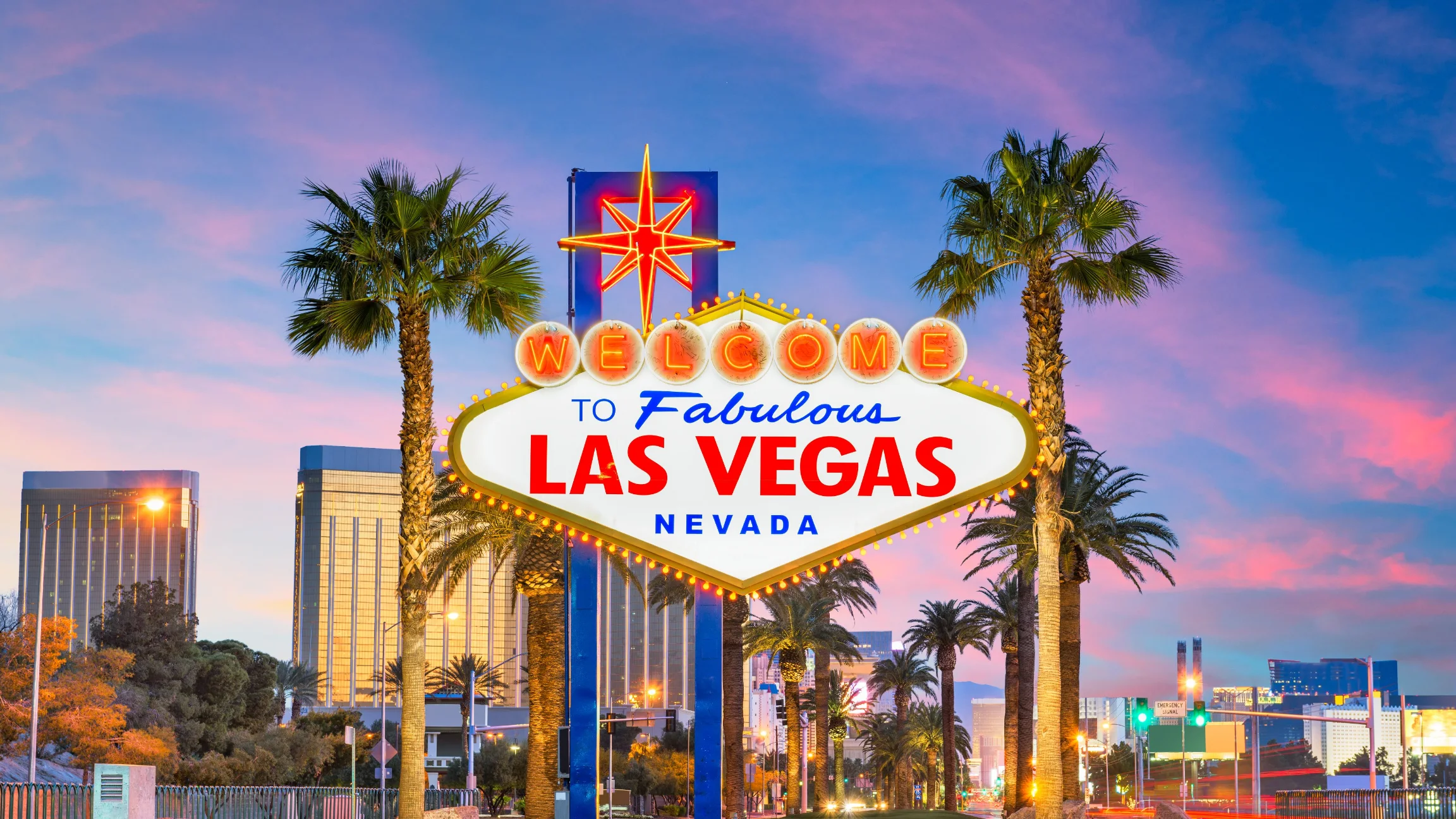 The iconic "Welcome to Fabulous Las Vegas, Nevada" sign is lit up at dusk, flanked by palm trees, with a backdrop of colorful city lights and a vibrant sky.