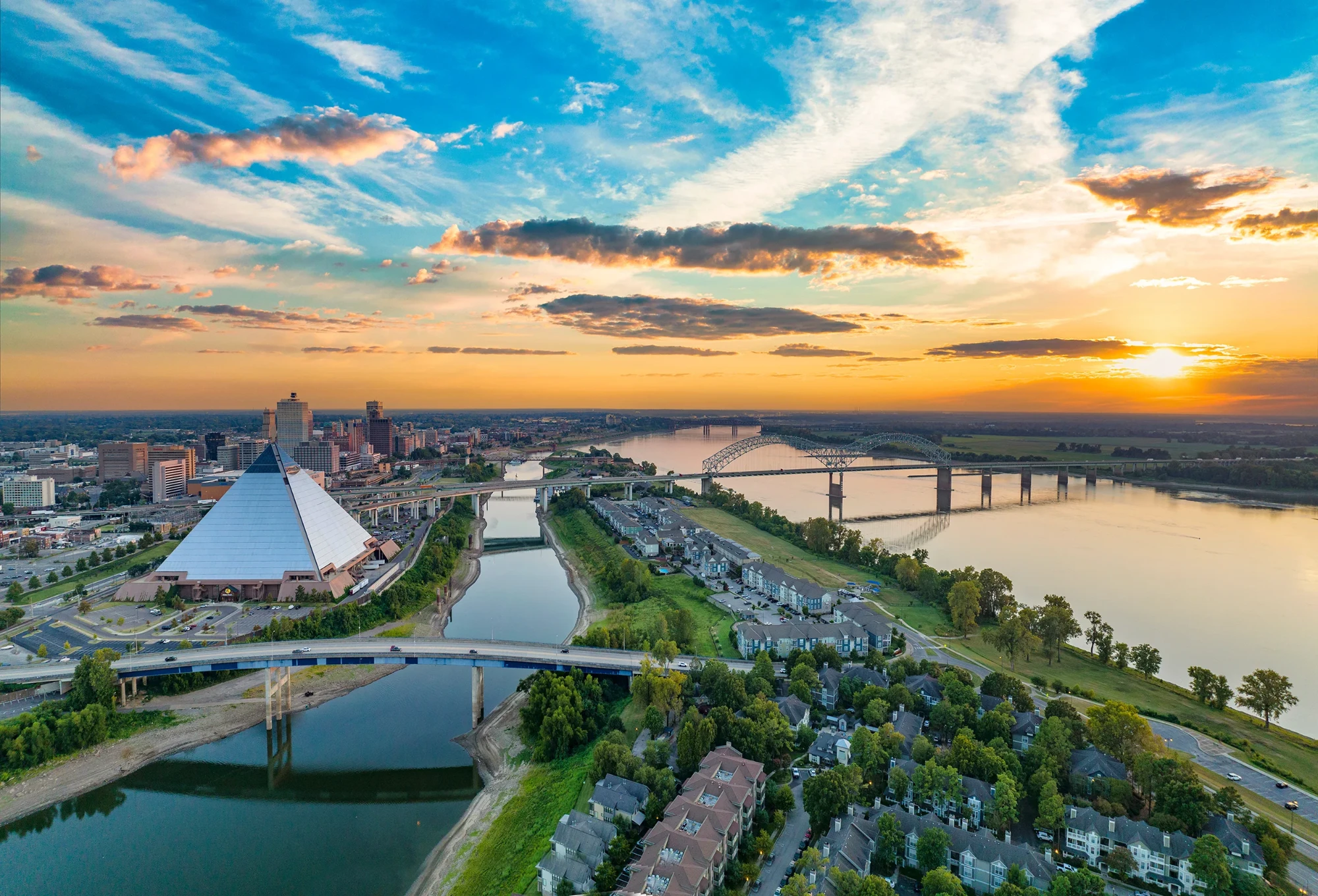 Aerial view of a cityscape at sunset, featuring a large glass pyramid on the left, a bridge over a river, residential areas, and a vibrant sky.