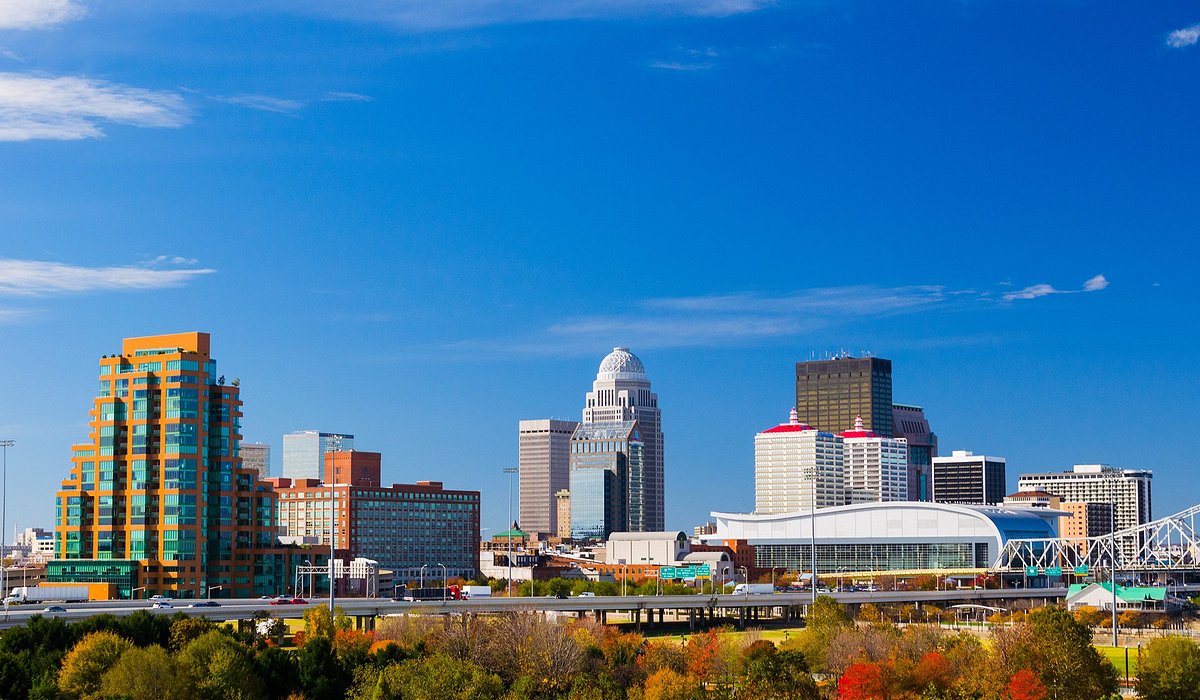 A vibrant city skyline under a clear blue sky, showcasing diverse architectural styles. Foreground features green and autumn-hued trees, conveying a lively urban atmosphere.