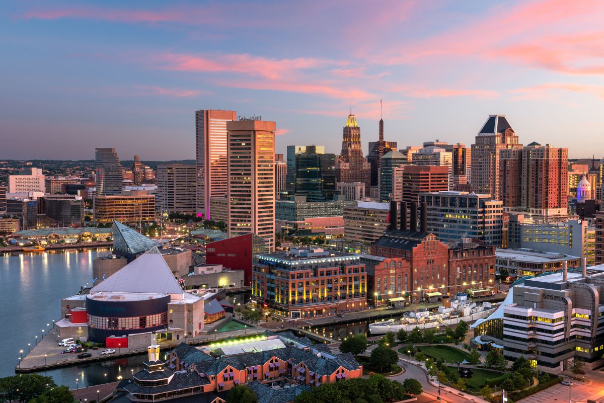 A vibrant cityscape of Baltimore at dusk, featuring illuminated buildings under a pink-purple sky. The harbor is visible, adding a serene waterfront element.