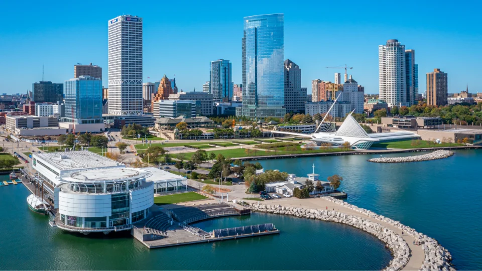 Aerial view of Milwaukee's downtown skyline with modern skyscrapers, a distinctive white pavilion, and a calm waterfront under a clear blue sky.