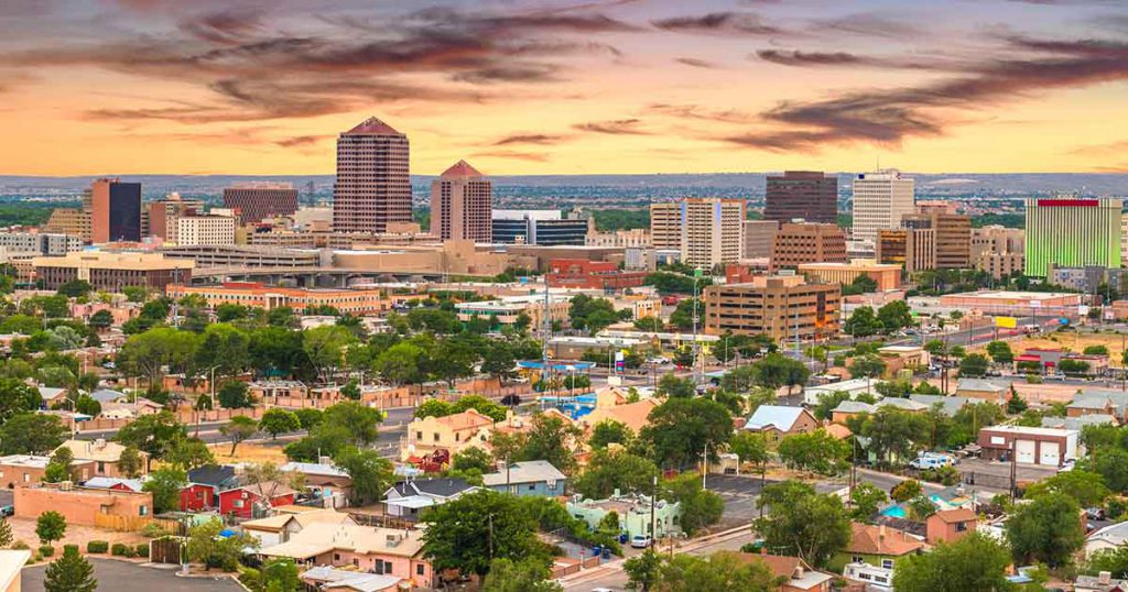Cityscape of Albuquerque at sunset, featuring a mix of modern skyscrapers and historic buildings. The sky is painted with warm hues, creating a serene atmosphere.