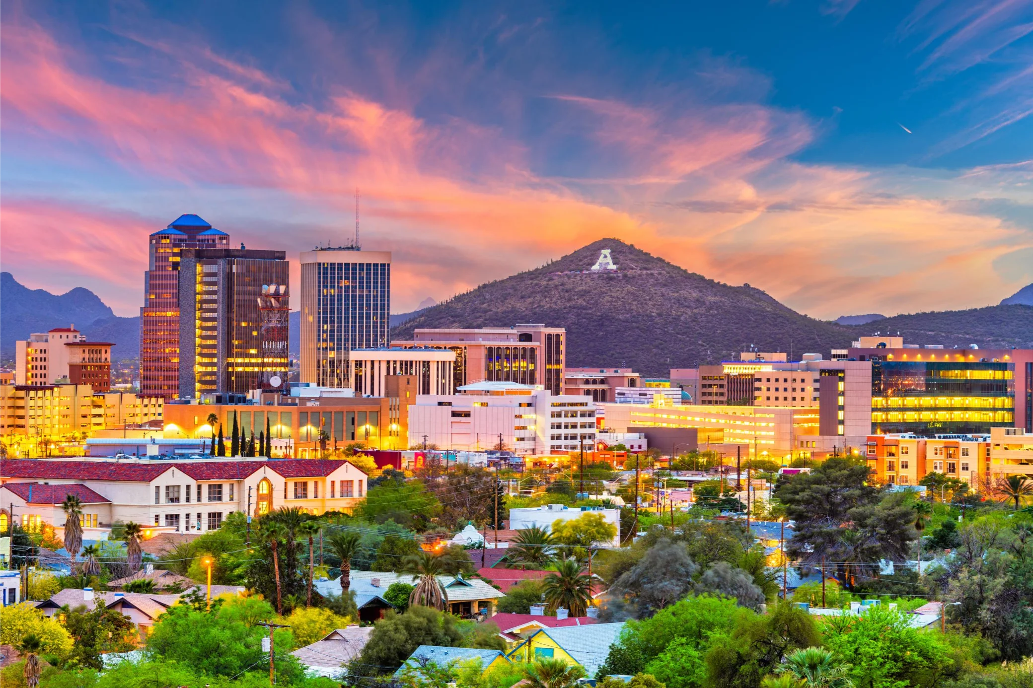 City skyline with modern buildings, lush greenery, and a mountain in the background at sunset. Vivid pink and orange clouds create a warm, vibrant atmosphere.