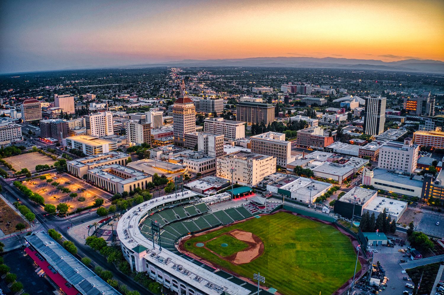 Aerial view of a cityscape at sunset, featuring a baseball stadium in the foreground. Tall buildings and a colorful sky create a serene atmosphere.