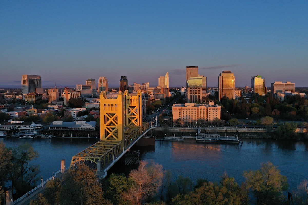 Aerial view of Sacramento at sunset, featuring the illuminated yellow Tower Bridge spanning the river. The city's skyline is bathed in warm, golden light.
