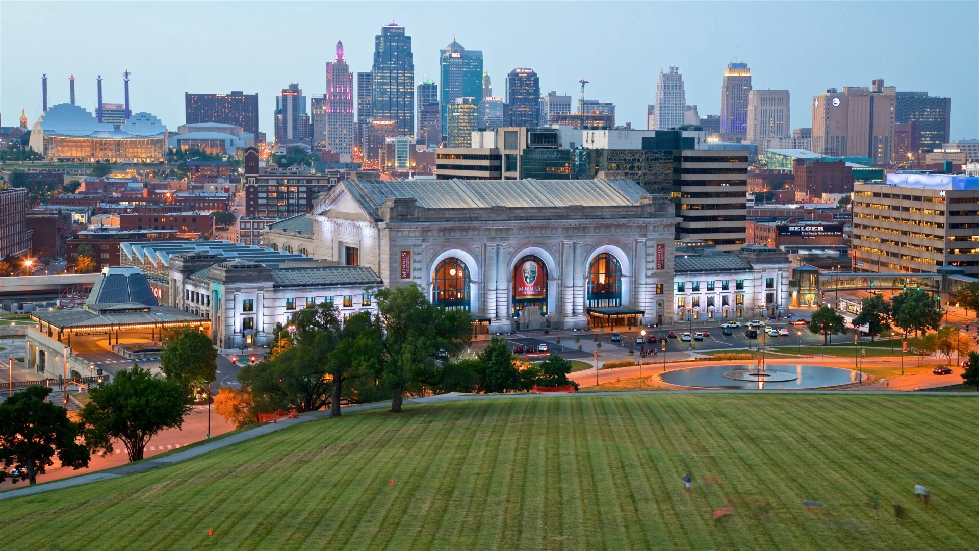 Aerial view of a historic train station with arched windows, set against a lit city skyline at dusk. Lush green lawn in the foreground.
