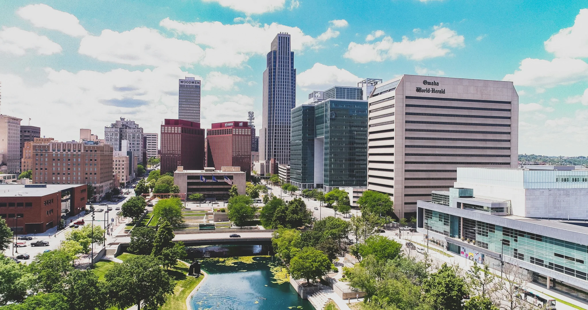 Aerial view of Omaha's skyline with modern skyscrapers, a mix of high-rise and historic buildings, lush green parks, and a tranquil pond under a partly cloudy sky.
