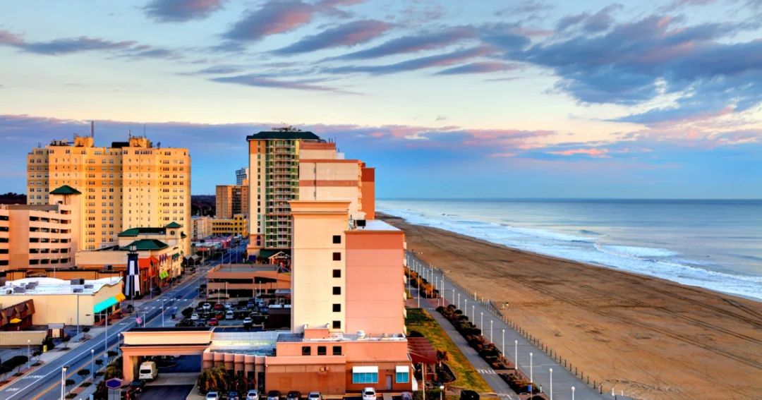 A coastal cityscape at sunset with tall buildings beside a sandy beach. Soft waves, pastel skies, and clouds create a serene, tranquil atmosphere.