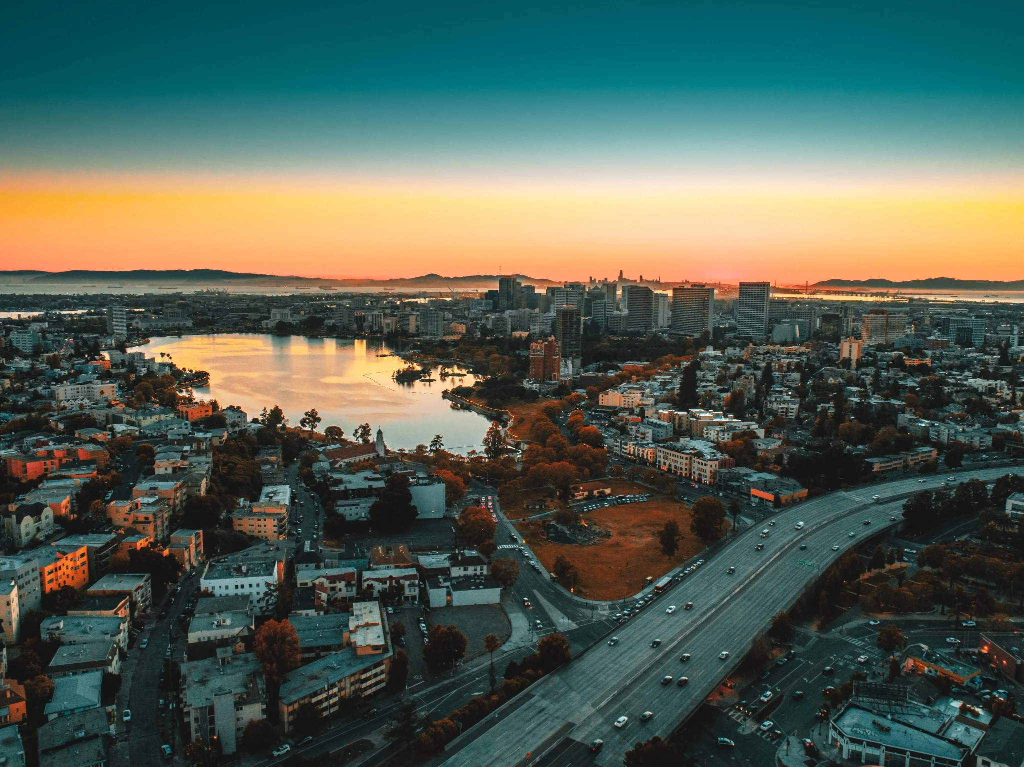 Aerial view of a cityscape at sunset, featuring a lake surrounded by urban buildings. The sky is a gradient of orange and blue, creating a serene vibe.