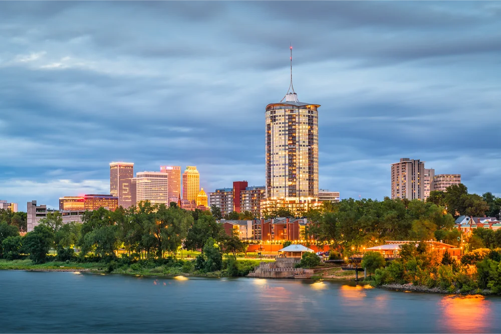 City skyline at dusk with tall, illuminated buildings against a cloudy sky. Trees line the waterfront, reflecting lights on the calm water.