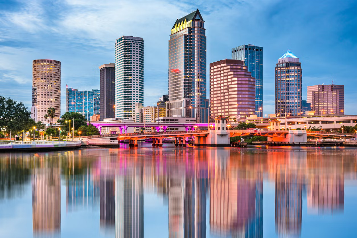A vibrant city skyline at dusk with tall buildings reflecting on calm waters. The scene is enhanced by colorful lights, creating a lively urban atmosphere.
