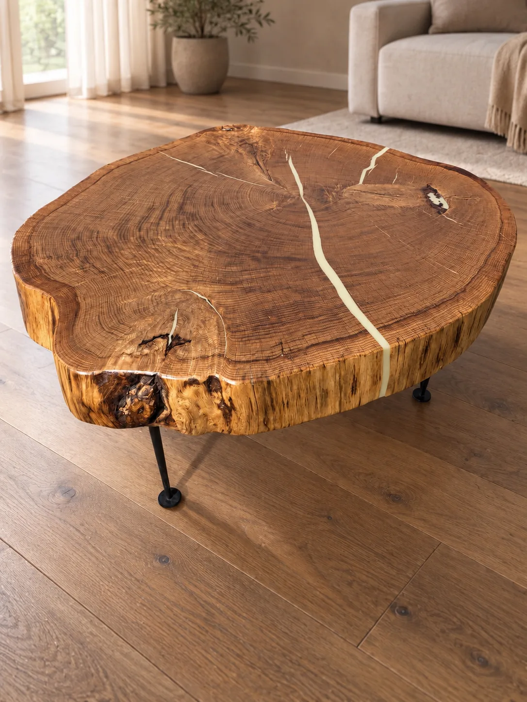 Moderner Couchtisch aus einer massiven, hellbraunen Eichenscheibe mit sichtbaren Jahresringen und natürlichen Rissen, die mit hellem Epoxidharz gefüllt sind, auf drei schwarzen Metallbeinen in einem hellen Wohnzimmer mit Holzboden, Pflanze und beige Sofaecke.