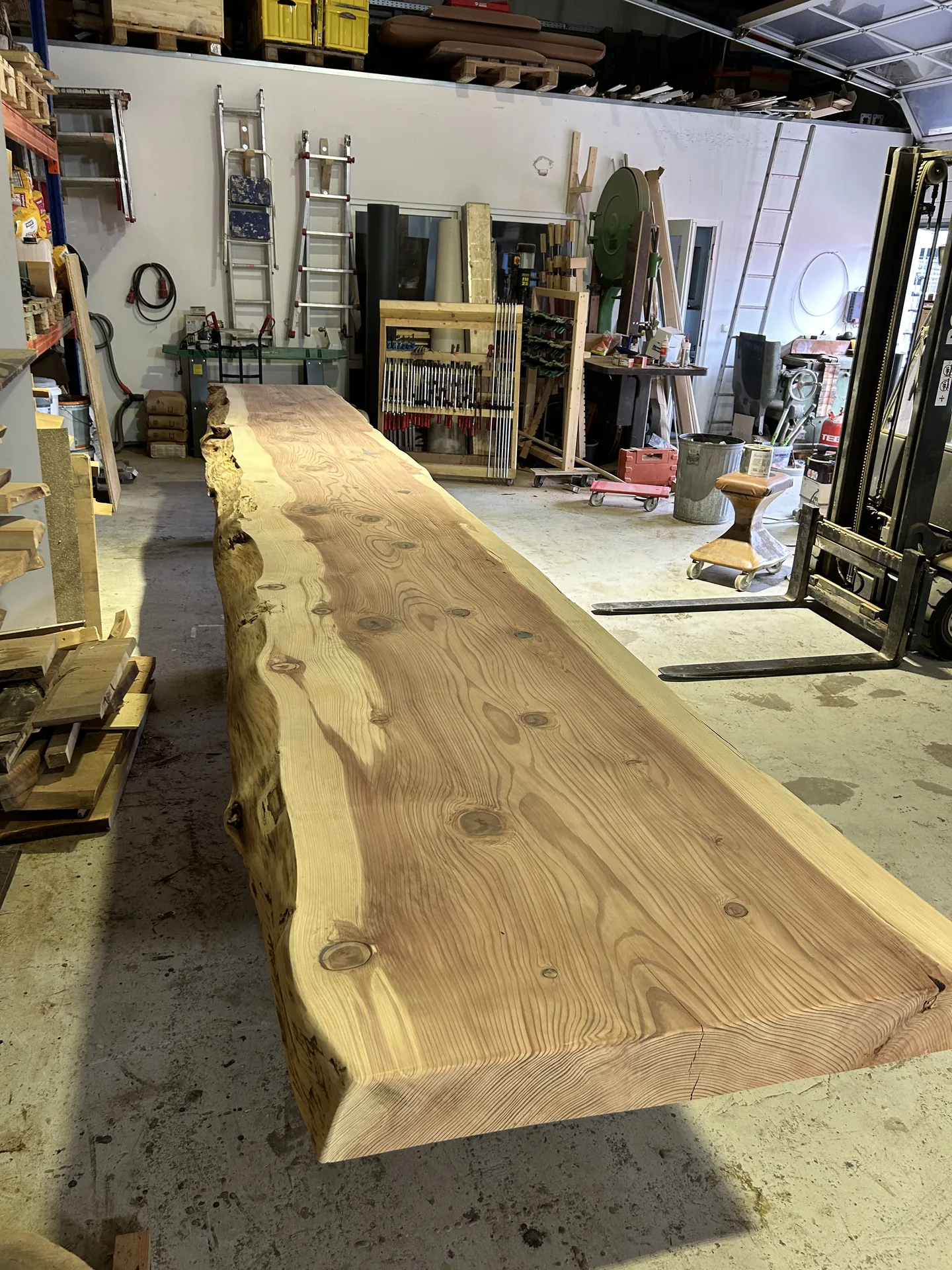 Lange, breite, naturbelassene Holzplatte mit sichtbarer Maserung und Baumkante in einer Werkstatt mit Werkzeugen, Leitern und einer Hebebühne im Hintergrund.