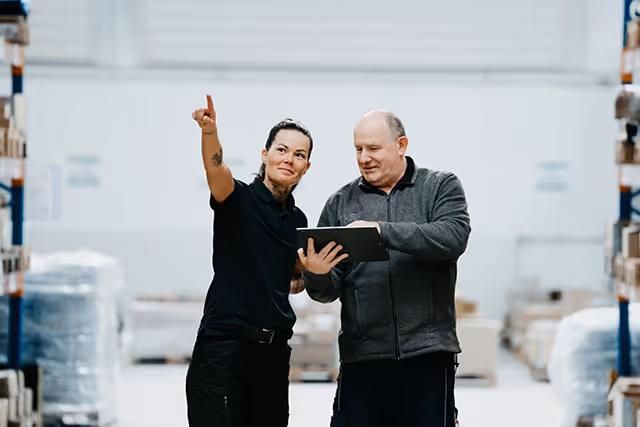 Zwei Mitarbeiter in einem Lager, eine Frau zeigt nach oben, während ein Mann ein Tablet hält.