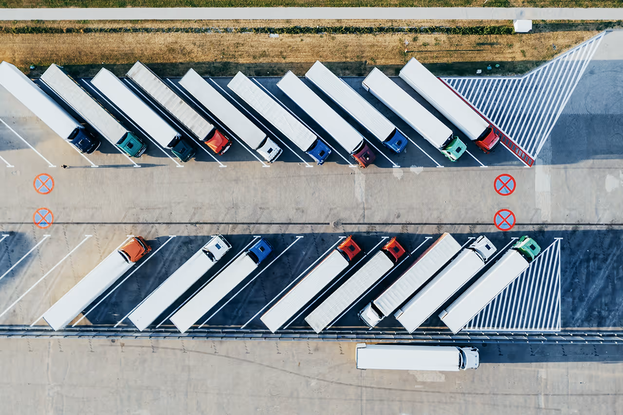 Luftaufnahme eines Parkplatzes mit Reihen von weißen Lastwagen auf beiden Seiten und einem einzelnen Lkw, der sich auf der Straße bewegt.