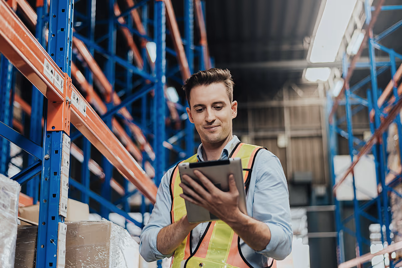 Mitarbeiter im Lager mit Warnweste überprüft Informationen auf einem Tablet vor Regalen mit Kartons.