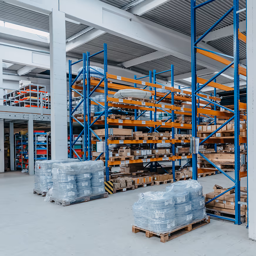 Lagerhalle mit blauen Metallregalen, gefüllt mit Kartons, und mehreren auf Paletten verpackten Waren.