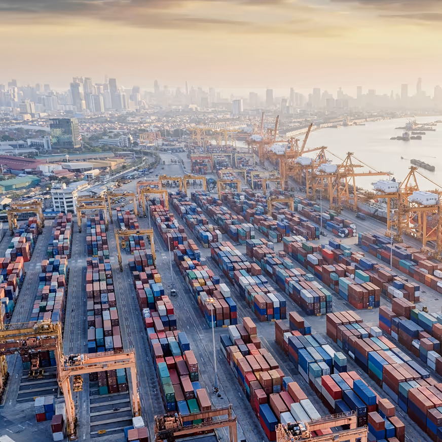 Luftaufnahme eines großen Seehafens mit unzähligen bunten Frachtcontainern und mehreren Kränen am Flussufer, dahinter eine neblige Stadtsilhouette bei Sonnenuntergang.