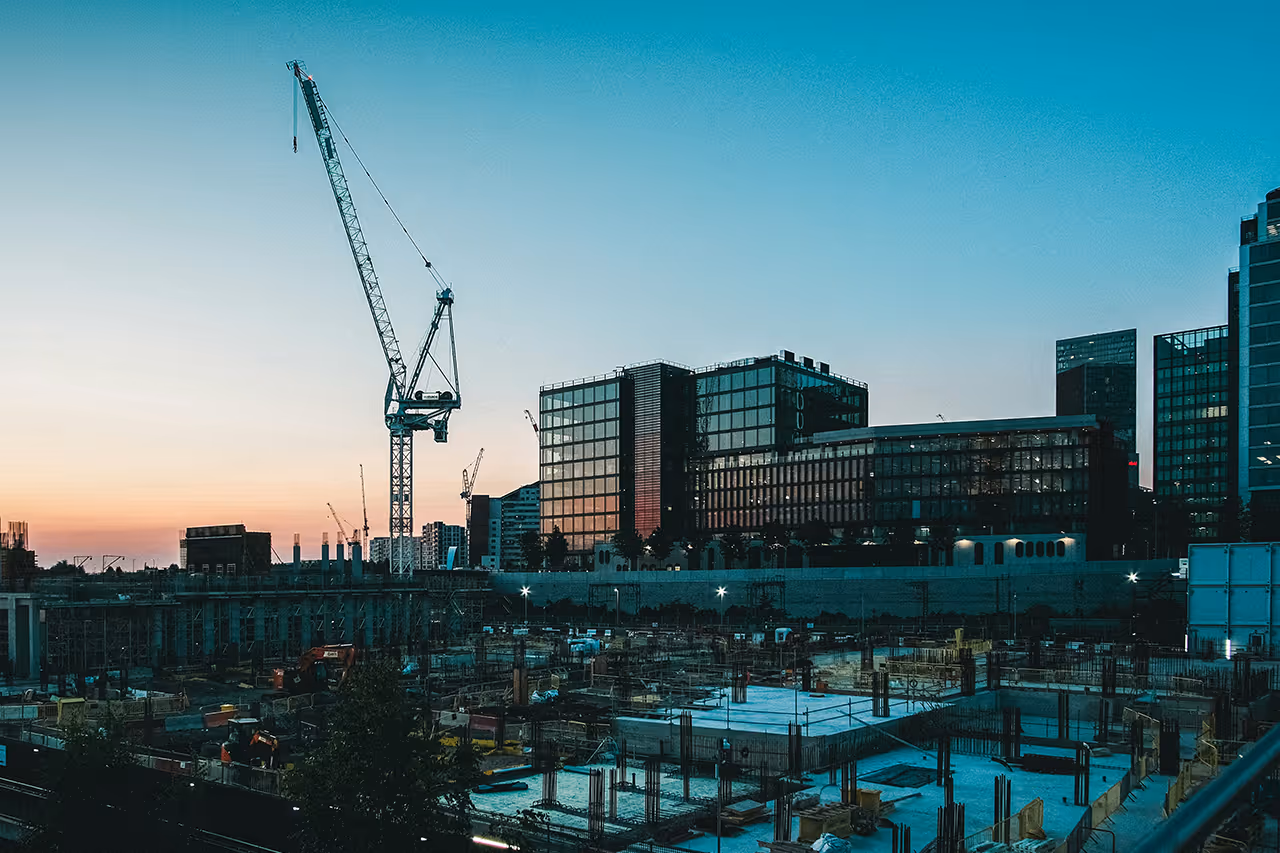 Baustelle mit Kran und mehreren Gebäuden im Hintergrund bei Dämmerung.