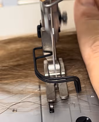 Une machine à coudre cousant des cheveux bruns, avec une main tenant l'aiguille en mouvement.