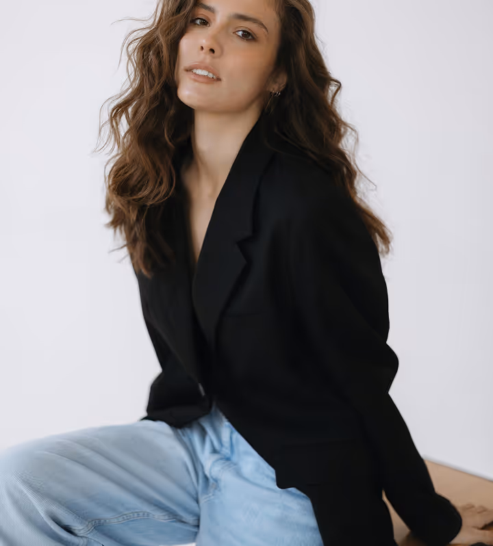 Jeune femme aux cheveux longs et bouclés portant une veste noire et un jean bleu clair, posant devant un fond blanc.
