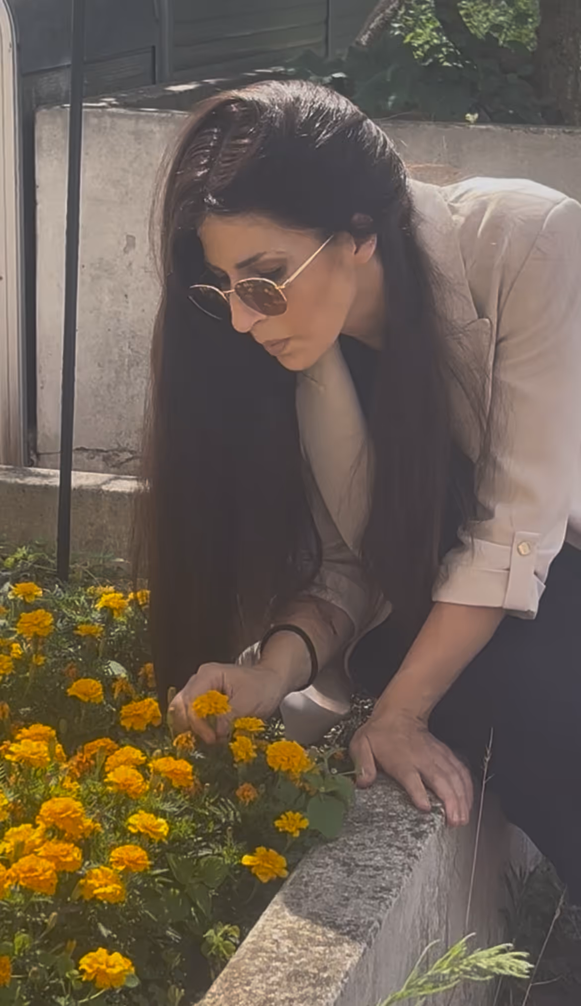 Femme aux cheveux longs et lunettes de soleil regardant et touchant des fleurs jaunes dans un jardin.