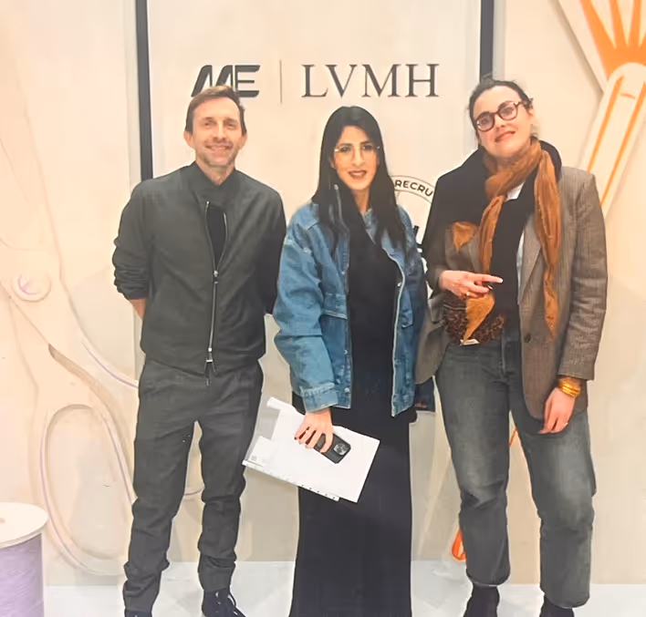 Trois personnes debout devant un mur avec le logo LVMH, une femme au centre porte une veste en jean et tient un carnet blanc.
