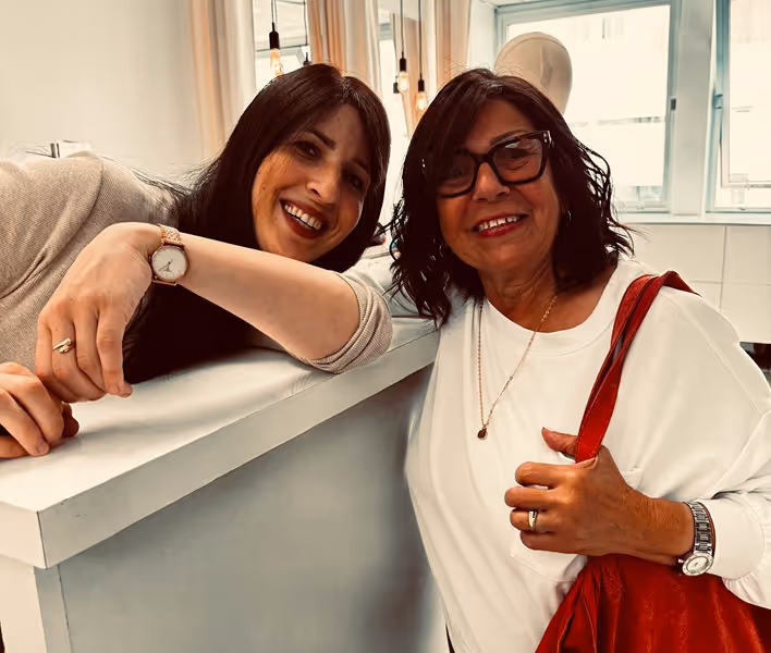 Deux femmes souriantes, une appuyée sur un comptoir blanc et l'autre portant un sac rouge et des lunettes.