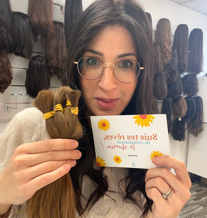 Une femme avec des lunettes rondes tient des touffes de cheveux attachés et une carte avec des motifs de tournesols dans un salon de perruques.