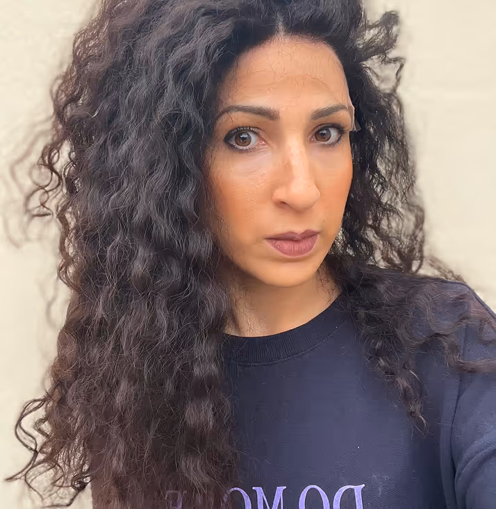 Portrait d'une femme aux longs cheveux bouclés foncés, portant un pull noir, regardant la caméra avec une expression neutre.