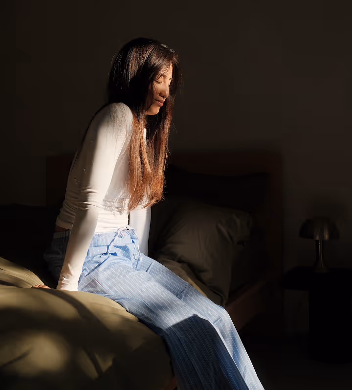 Femme assise sur un lit, vêtue d'un haut blanc et d'un pantalon rayé bleu clair, illuminée par la lumière du soleil dans une chambre sombre.