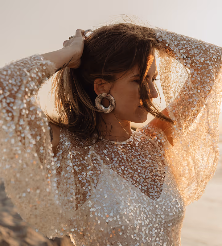 Femme aux cheveux bruns relevés, portant une blouse transparente à sequins et de grandes boucles d'oreilles rondes, éclairée par la lumière douce du coucher de soleil.