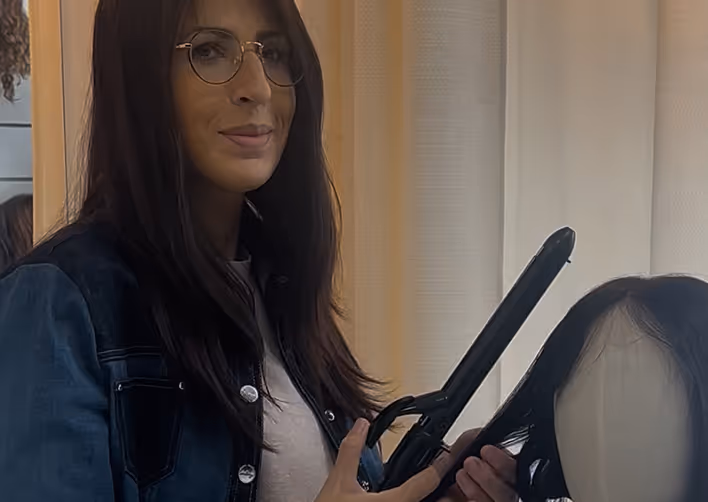 Une femme avec des lunettes utilise un fer à lisser pour coiffer une perruque noire sur une tête de mannequin.