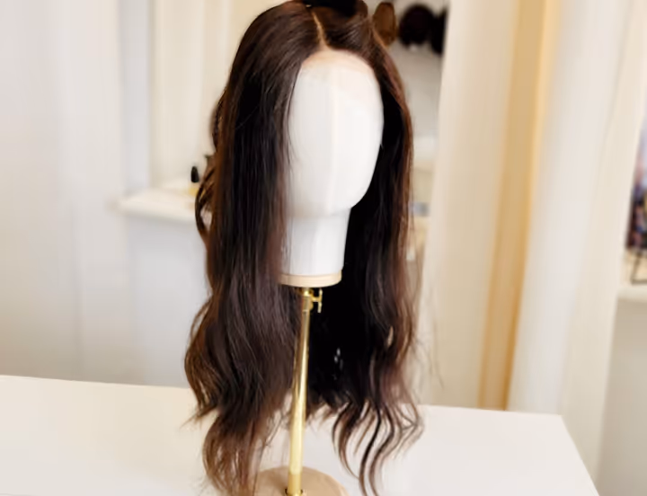 Perruque brune aux cheveux longs ondulés exposée sur un support blanc et doré.