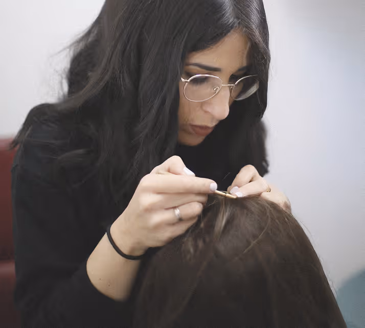 Une femme aux cheveux noirs et lunettes pose délicatement une extension ou un accessoire capillaire sur les cheveux d'une autre personne.
