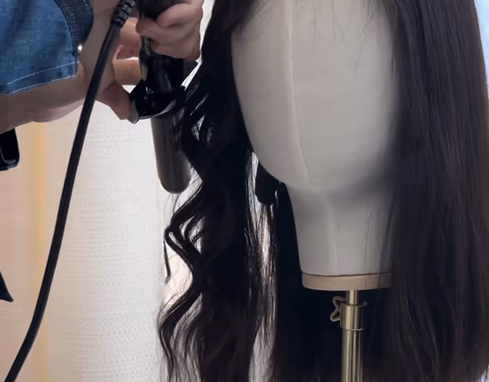 Une main utilise un fer à friser pour boucler une mèche de cheveux noirs sur une tête à perruque en toile beige.