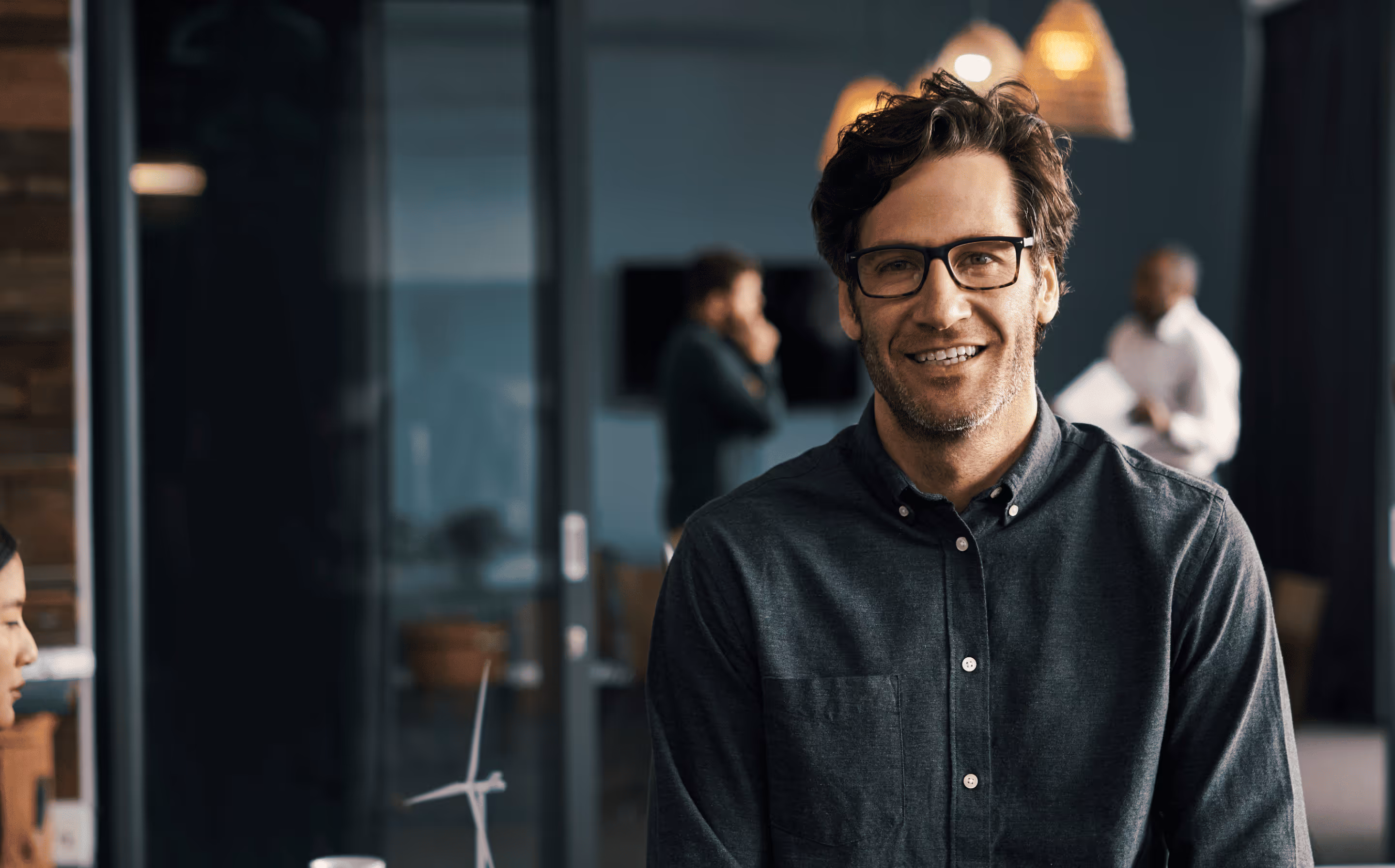 Smiling man with glasses and a dark button-up shirt in a modern office with coworkers in the background.