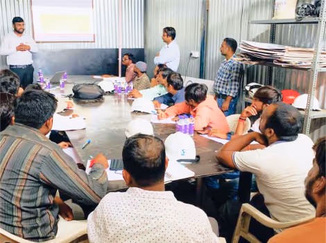 Group of men attentively listening to a presenter in a metal-walled training room with a large table and documents.