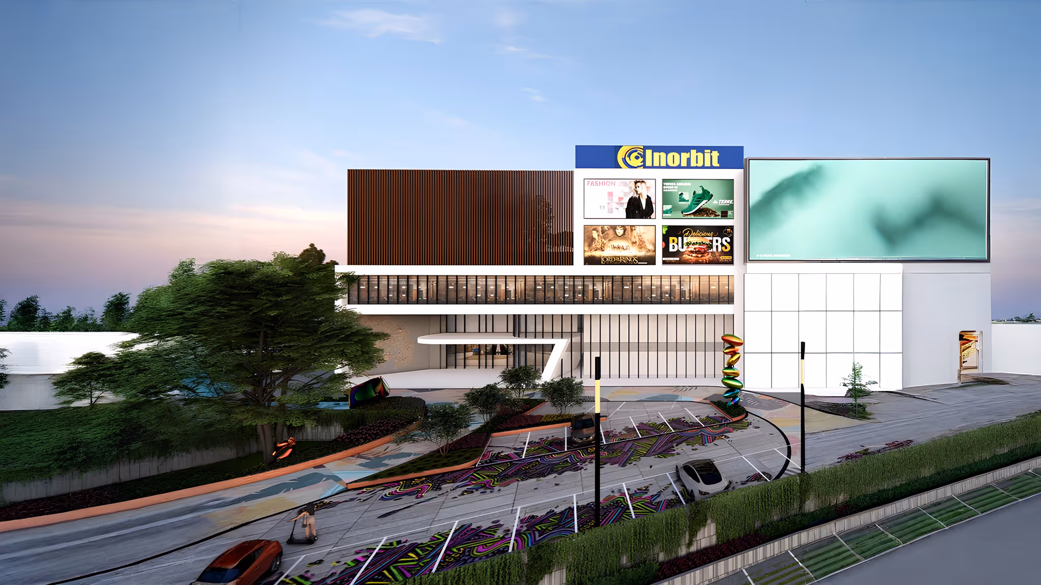 Modern commercial building with large digital billboards, colorful decorative pavement, and surrounding greenery under a clear sky.