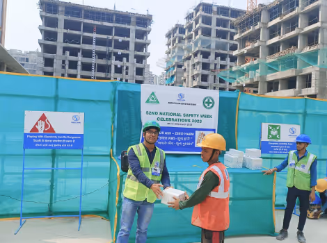 Construction workers wearing helmets and safety vests at a building site distributing safety gear with banners promoting National Safety Week in the background.