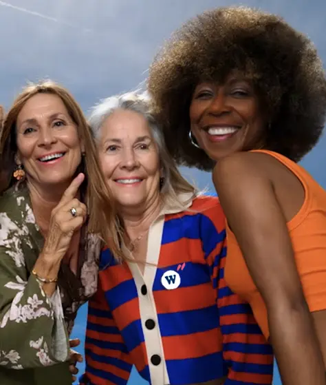 Trois femmes souriantes de différents âges et origines posant ensemble devant un ciel bleu.
