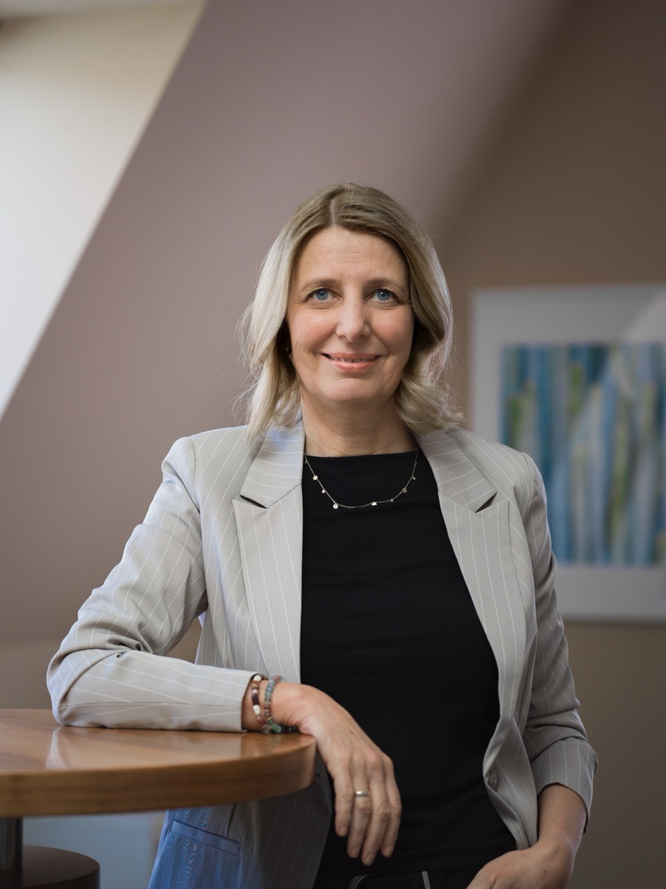 Smiling middle-aged woman with blonde hair wearing a gray pinstripe blazer and black top sitting at a wooden table indoors.