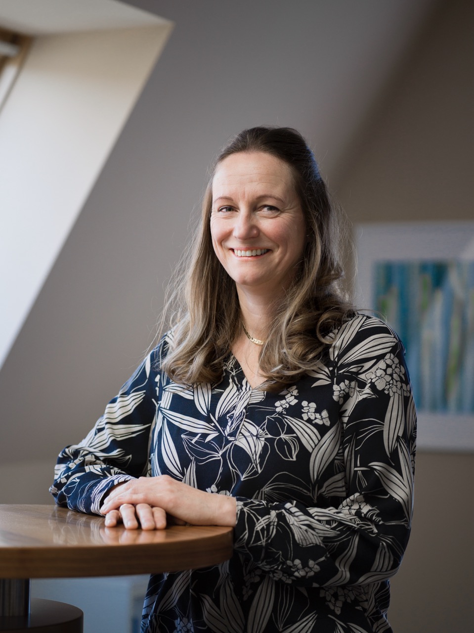 Smiling woman with long wavy hair wearing a black and white floral blouse, sitting at a wooden table in a softly lit room.