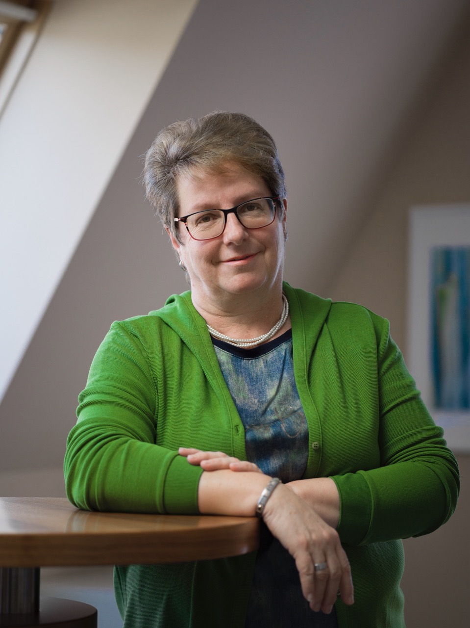 Middle-aged woman with short hair and glasses wearing a green cardigan and pearl necklace, sitting at a wooden table with hands folded.