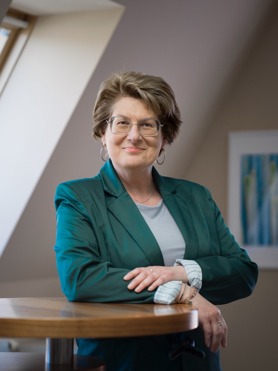 Woman with short hair and glasses wearing a green blazer, leaning on a wooden table with arms crossed, smiling.