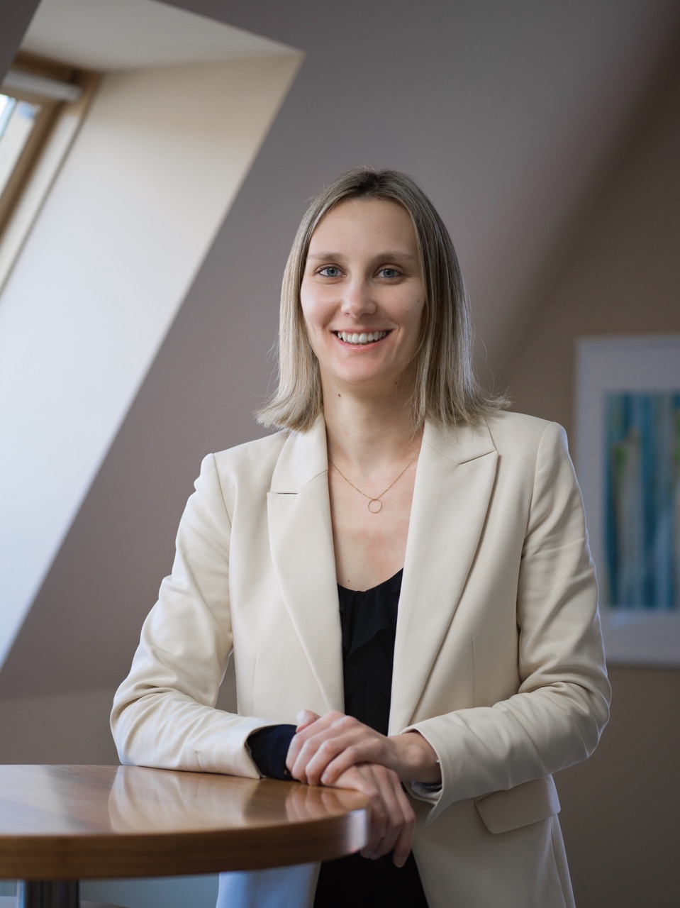 Smiling woman with blonde hair wearing a beige blazer and black top, standing behind a wooden table in a softly lit room.